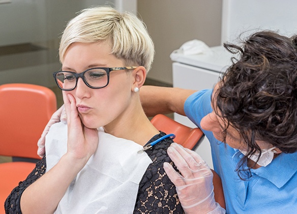 A young woman wearing glasses and holding her cheek in pain while a dentist prepares to help remedy the problem