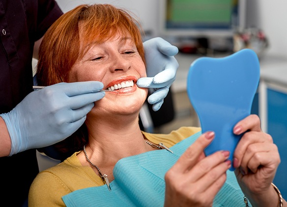 A middle-aged woman with red hair admiring her new smile in the mirror while at the dentist’s office